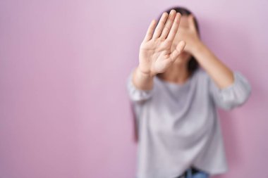 Young brunette woman standing over pink background covering eyes with hands and doing stop gesture with sad and fear expression. embarrassed and negative concept. 