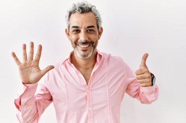 Orta yaşlı İspanyol adam izole edilmiş arka planda duruyor. Altıncı parmaklarını gösterip işaret ederken kendinden emin ve mutlu bir şekilde gülümsüyor.. 