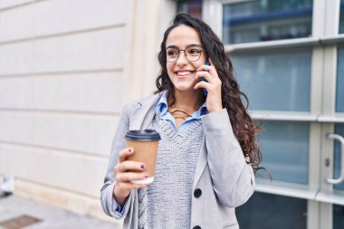Genç İspanyol kadın yönetici kahve içiyor ve caddede akıllı telefondan konuşuyor.