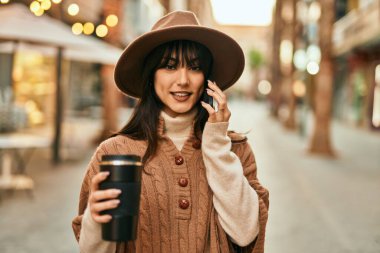 Kışlık şapkalı esmer kadın telefonda konuşuyor ve şehirde bir fincan kahve içiyor.