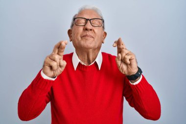 Senior man with grey hair standing over isolated background gesturing finger crossed smiling with hope and eyes closed. luck and superstitious concept. 