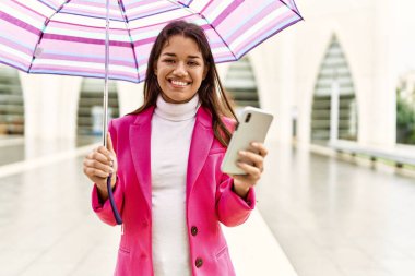 Young latin woman using smartphone holding umbrella at street