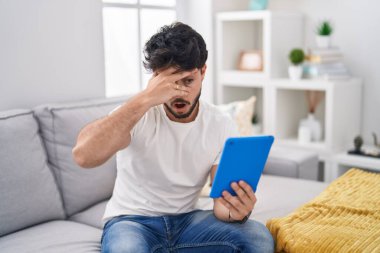 Hispanic man with beard using touchpad sitting on the sofa peeking in shock covering face and eyes with hand, looking through fingers with embarrassed expression. 