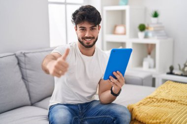Hispanic man with beard using touchpad sitting on the sofa approving doing positive gesture with hand, thumbs up smiling and happy for success. winner gesture. 