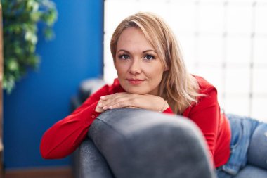 Sarışın genç bir kadın evdeki koltukta kendinden emin bir şekilde oturarak gülümsüyor.