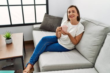 Orta yaşlı İspanyol kadın kanepede oturuyor ve evde bir fincan kahve içiyor.