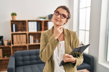 Muayenehanede çalışan genç bir kadın elleri çenesinde soru sorarken, düşünceli bir ifade takınıyor. Düşünceli bir yüzle gülümsüyor. şüphe kavramı. 