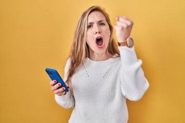 Genç sarışın kadın akıllı telefon kullanarak kızgın ve kızgın bir şekilde mesaj yazıyor. Öfkeyle bağırırken yumruğunu kaldırıyor ve öfkeleniyor. öfke ve agresif konsept. 