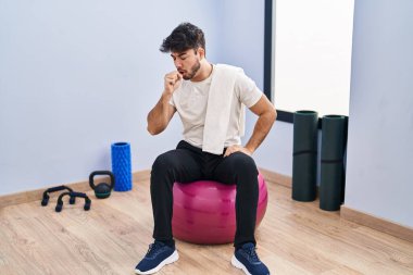 Hispanic man with beard sitting on pilate balls at yoga room feeling unwell and coughing as symptom for cold or bronchitis. health care concept. 