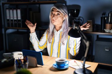 Gri saçlı, geceleri ofiste çalışan, kafası karışmış, elleri ve kolları havada orta yaşlı bir kadın. şüphe kavramı. 
