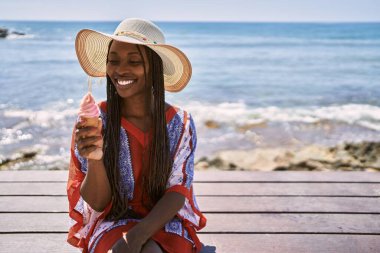 Young african american woman eating ice cream sitting on the bench at the beach.