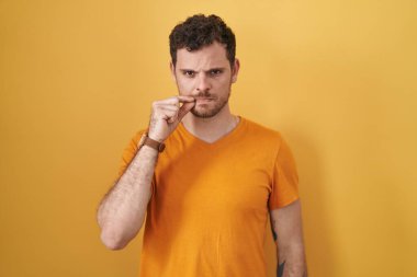 Sarı arka planda dikilen İspanyol genç adam ağzı ve dudakları parmaklarıyla kapanmış. gizli ve sessiz, tabu konuşması 
