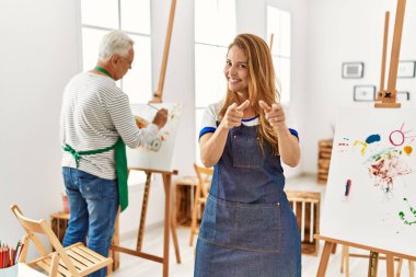 Hispanic woman wearing apron at art studio pointing fingers to camera with happy and funny face. good energy and vibes. 