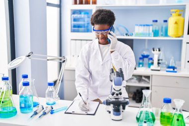 Bilim adamı üniforması giyen Afrikalı Amerikalı kadın. Akıllı telefondan konuşuyor. Laboratuvarda panoya yazı yazıyor.