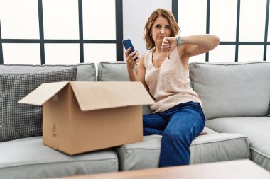Orta yaşlı bir kadın kanepede oturmuş telefondaki teslimat paketini kontrol ediyor. Kızgın suratlı, olumsuz işaretli. Başparmakları aşağıda, reddedilme kavramından hoşlanmadığını gösteriyor. 