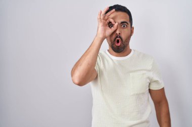 Hispanic man with beard standing over isolated background doing ok gesture shocked with surprised face, eye looking through fingers. unbelieving expression. 