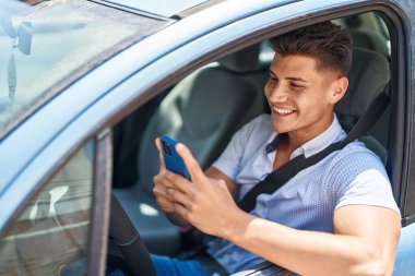 Young hispanic man using smartphone sitting on car at street
