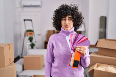 Yeni ev duvarının rengini seçen kıvırcık saçlı, esmer, genç bir kadın. Düşünme tavrı ve kendine güvenen ayık bir ifade. 