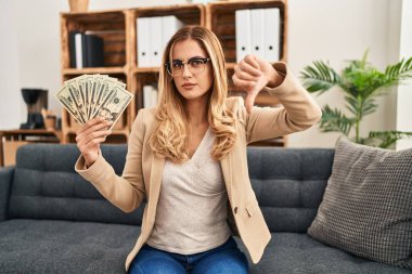 Terapi ofisinde çalışan genç sarışın kadın elinde kızgın yüzle para tutuyor. Negatif işaret baş parmakları aşağıda, reddedilme konsepti ise... 