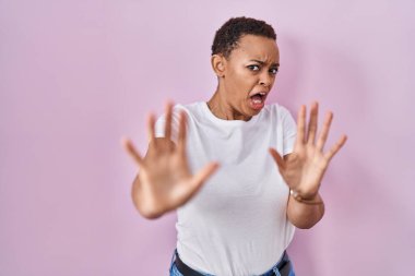 Güzel afro-amerikan kadın pembe arka planda duruyor korku ifadesinden korkuyor ve dehşete düşüyor jesti elleriyle durdurun, şok içinde bağırıyor. panik konsepti. 