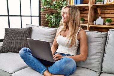 Evdeki kanepede dizüstü bilgisayar kullanan genç sarışın kadın.