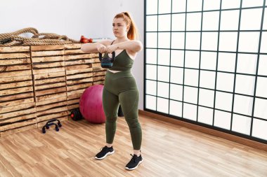 Genç kızıl saçlı kadın spor merkezinde kettlebell ile antrenman yapıyor.