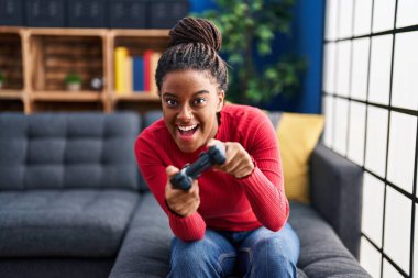 Evdeki kanepede oturan Afrikalı Amerikalı kadın video oyunu oynuyor.