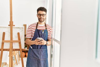 Sanat stüdyosunda akıllı telefon kullanarak gülümseyen genç Arap sanatçı..