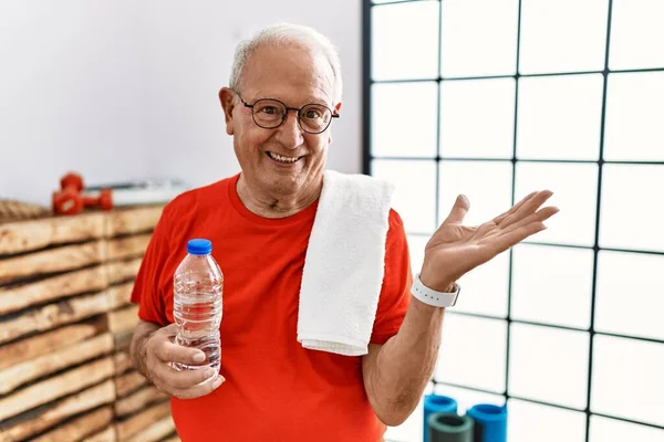 Senior man wearing sportswear and towel at the gym smiling cheerful presenting and pointing with palm of hand looking at the camera.