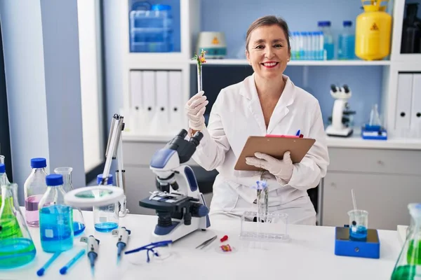 Laboratuvarda elinde çiçek okuma panosuyla test tüpü tutan orta yaşlı bir bilim kadını.