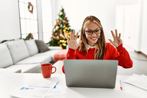 年轻的高加索女孩坐在桌子上 用笔记本电脑在圣诞树旁工作 展示并用9号手指指尖 同时带着自信和快乐的笑容 — 图库照片