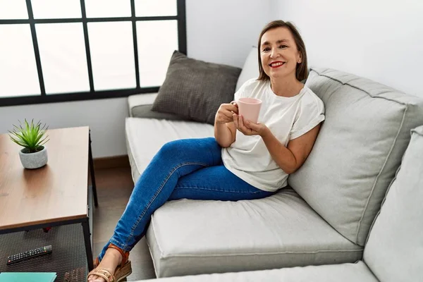 Orta yaşlı İspanyol kadın kanepede oturuyor ve evde bir fincan kahve içiyor.