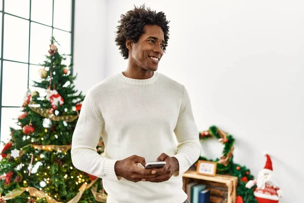 Young african american man uisng smartphone standing by christmas tree at home.