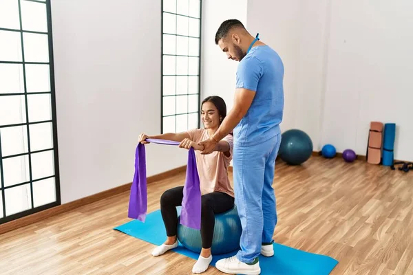 Latin man and woman wearing physiotherapist uniform having rehab session using fit ball and elastic band at rehab center