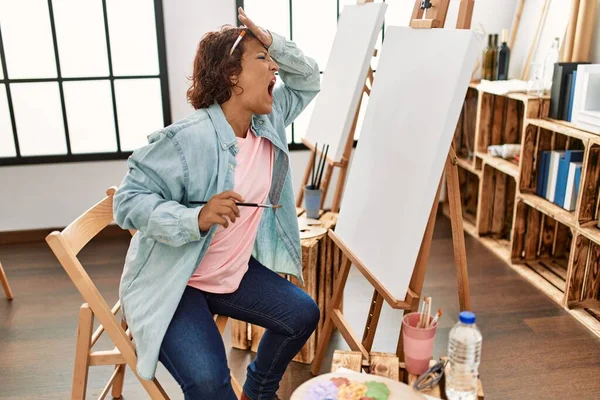 Middle age hispanic woman at art studio painting on canvas stressed and frustrated with hand on head, surprised and angry face