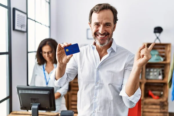 Hispanic man holding credit card at retail shop smiling happy pointing with hand and finger to the side