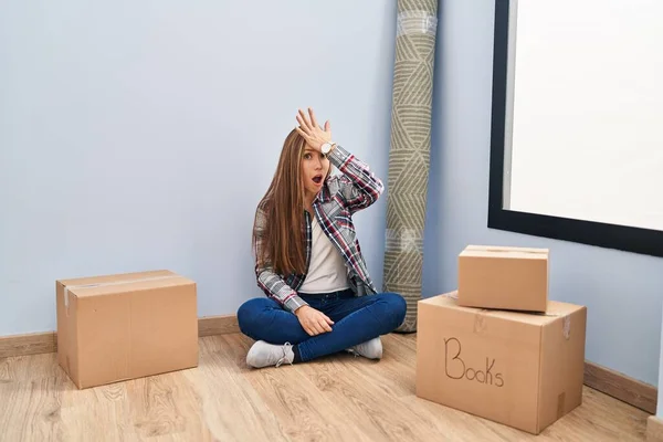 Young blonde woman sitting on the floor moving to a new home surprised with hand on head for mistake, remember error. forgot, bad memory concept.