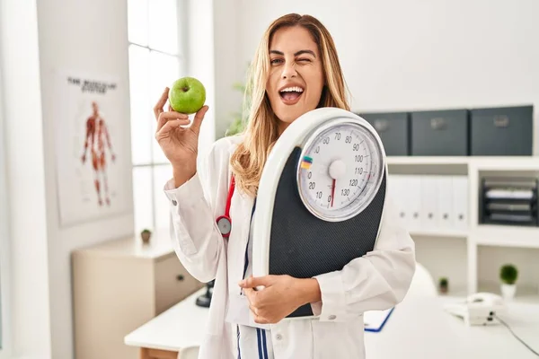 年轻的金发碧眼的医生女人拿着称重的机器 绿色的苹果眨着眼睛 带着性感的表情 笑容满面地看着摄像机 — 图库照片