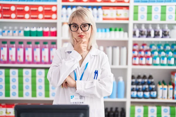 Young caucasian woman working at pharmacy drugstore looking confident at the camera with smile with crossed arms and hand raised on chin. thinking positive.