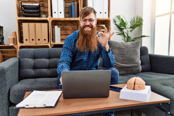 红头发的男人留着长胡子 用笔记本电脑在心理诊所工作 用手指做手势 微笑友善的手势 展示出极好的象征 — 图库照片