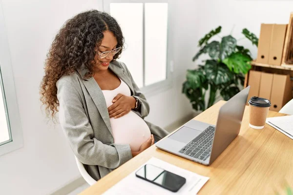 年轻的拉丁女人怀孕了 带着自信的笑容摸着肚子在办公室工作 — 图库照片