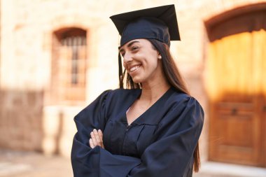 Young hispanic woman wearing graduated uniform standing with arms crossed gesture at university