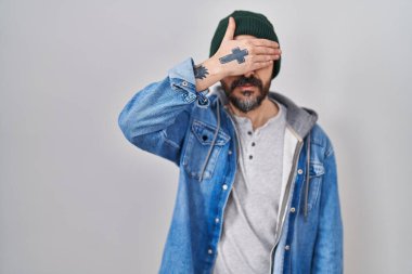Young hispanic man with tattoos wearing wool cap smiling and laughing with hand on face covering eyes for surprise. blind concept. 