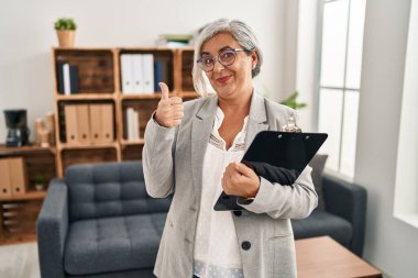 Middle age woman with grey hair at consultation office doing happy thumbs up gesture with hand. approving expression looking at the camera showing success. 