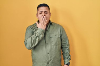 Hispanic young man standing over yellow background bored yawning tired covering mouth with hand. restless and sleepiness. 