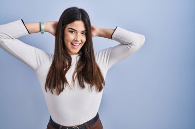 Genç esmer kadın mavi arka planda duruyor rahatlıyor ve esniyor, kolları ve elleri başının arkasında mutlu bir şekilde gülümsüyor. 