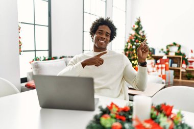 Genç Afrikalı Amerikalı adam dizüstü bilgisayar kullanıyor Noel ağacının yanında oturuyor gülümsüyor ve kameraya bakıyor iki eliyle ve parmaklarıyla yan tarafa bakıyor.. 