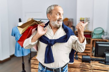 Butik dükkanında alışveriş torbaları tutan yakışıklı bir adam başparmağıyla yan tarafı işaret ediyor ve ağzı açık gülümsüyor. 