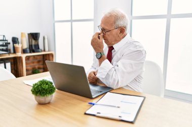 Senior man working at the office using computer laptop tired rubbing nose and eyes feeling fatigue and headache. stress and frustration concept. 