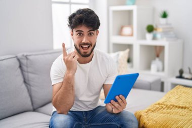 Hispanic man with beard using touchpad sitting on the sofa pointing finger up with successful idea. exited and happy. number one. 
